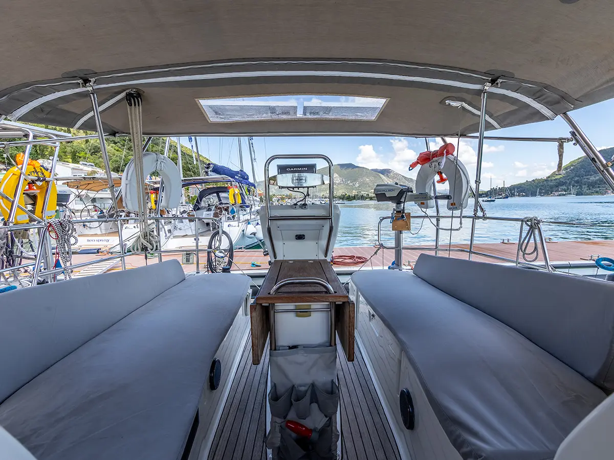 View from the rear deck of a Bavaria 36 yacht with cushioned seats looking out towards a harbor filled with boats and a lush green coastline under a clear blue sky.