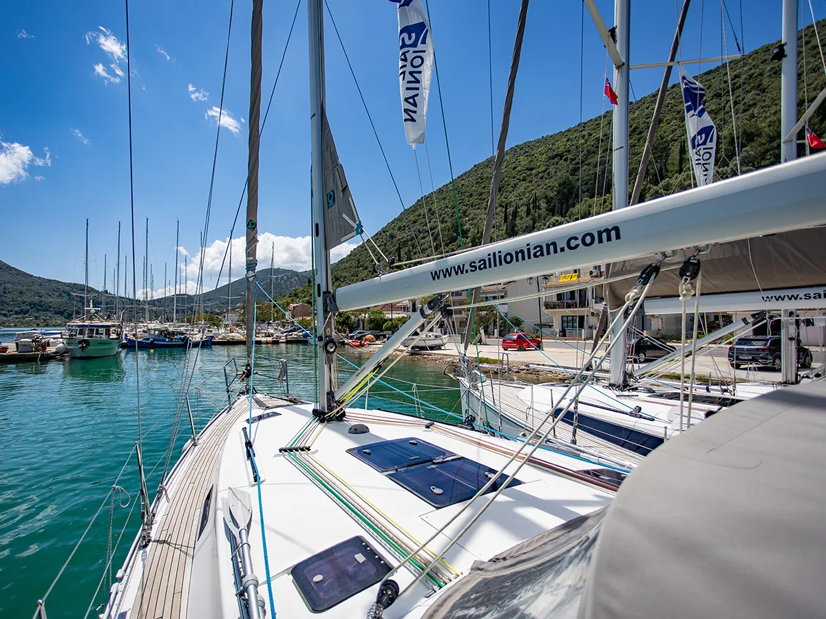 View from the deck of a Bavaria 36 sailboat in a marina with multiple yachts moored, surrounded by lush green hills under a clear blue sky. A website banner reading "sailionian.com"