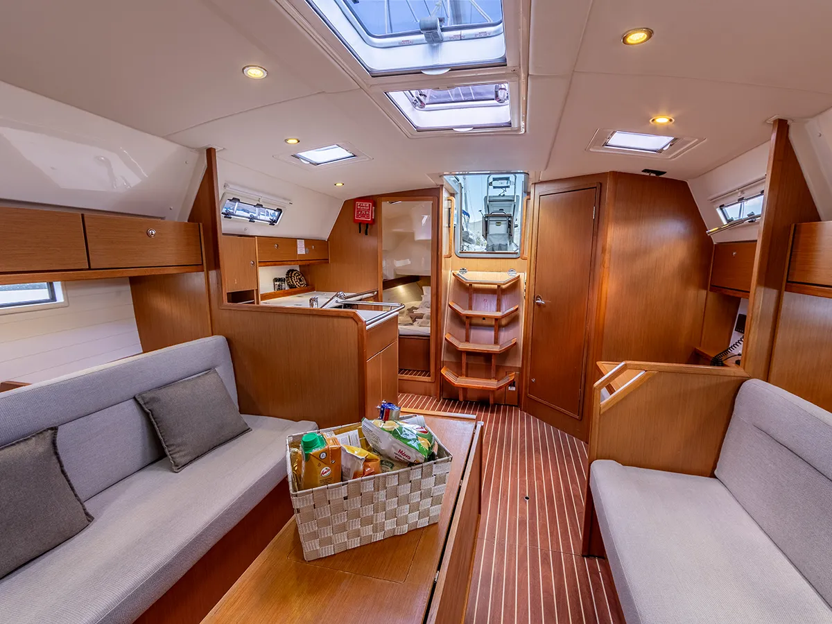 Interior view of a Bavaria 36 sailboat cabin featuring a cozy seating area with light gray cushions, a small kitchen area in the background, and polished wooden flooring and cabinets. A basket of groceries