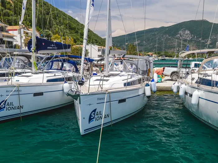 Several white sailboats, including a Bavaria 37 Charter, are moored side by side in clear turquoise water at a marina, with green hills and buildings in the background and 