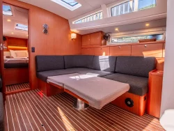 The interior of this Bavaria Vision 42 features a spacious living area with a large gray L-shaped sofa and a foldable table. Wooden paneling and striped flooring add a warm touch, while sunlight filters through multiple skylights and windows, illuminating the modern and cozy space.