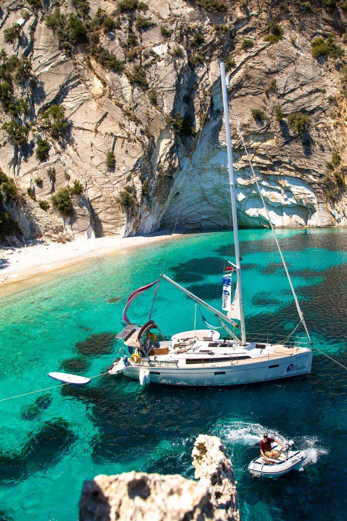 A Bavaria 37 sailboat anchored in a clear turquoise bay near steep rocky cliffs, with a dinghy approaching it under a bright sun.