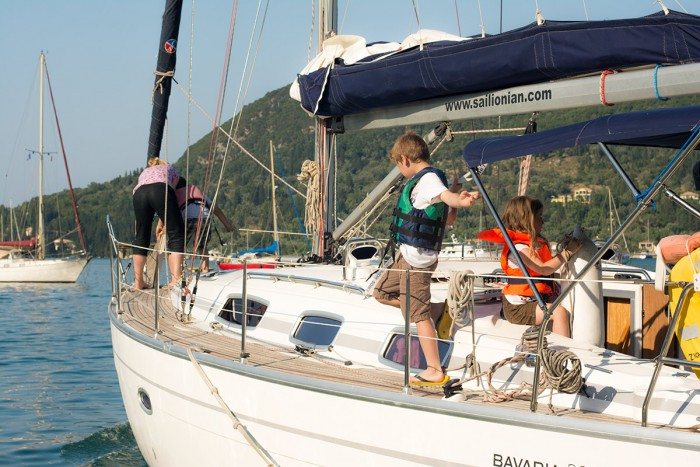 Family prepares on a Sail Ionian Bavaria 37 sailboat at a marina with lush hills in the background; a child in a life jacket interacts with an adult, another stands by the rail. Learn to sail in Greece.