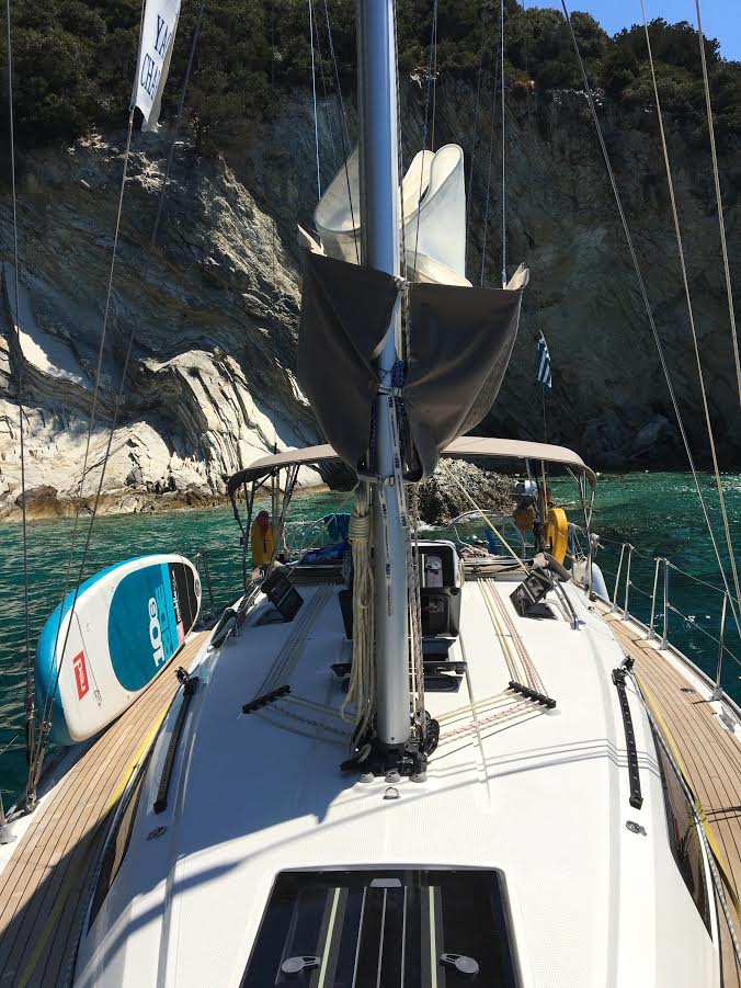A Bavaria 37 sailboat moored near a rocky shore with clear blue water, viewed from the deck looking forward towards the furled sails and surrounding cliffs.