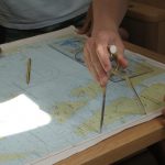 Hands of two people using a divider tool on a nautical map, with pencils and a compass, planning a yachtmaster theory route.
