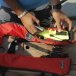 A person on a boat prepares a red life jacket with a yellow whistle attached during an RYA training course. The focus is on their hands as they adjust the jacket on a wooden surface.