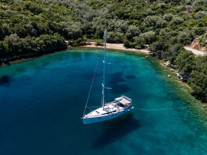 A Bavaria 46 Style sailboat anchored in a secluded, clear turquoise bay surrounded by lush green forest. The shoreline is a mix of sandy beach and rocky terrain, creating a peaceful and picturesque scene ideal for relaxation.