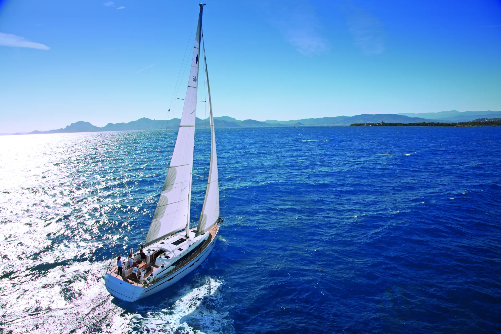 A white Bavaria 46 Style sailboat with people aboard sails on bright blue water under a clear sky, with distant mountains visible on the horizon.