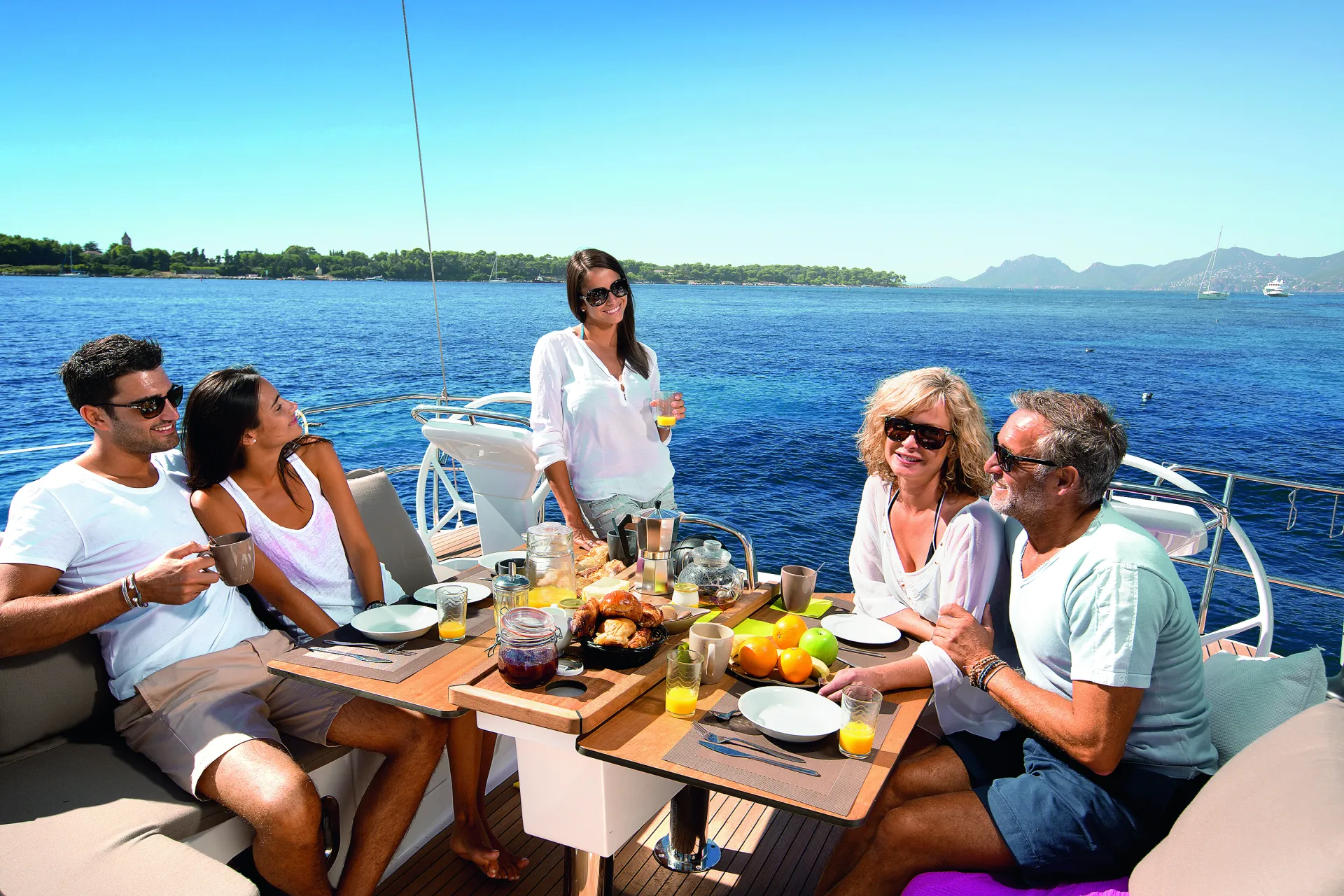 Five people enjoy a meal and drinks on a Bavaria 46 Style yacht with a scenic sea view, blue skies, and a distant shoreline. The table is set with fruit, pastries, and juice, and everyone appears relaxed and happy.