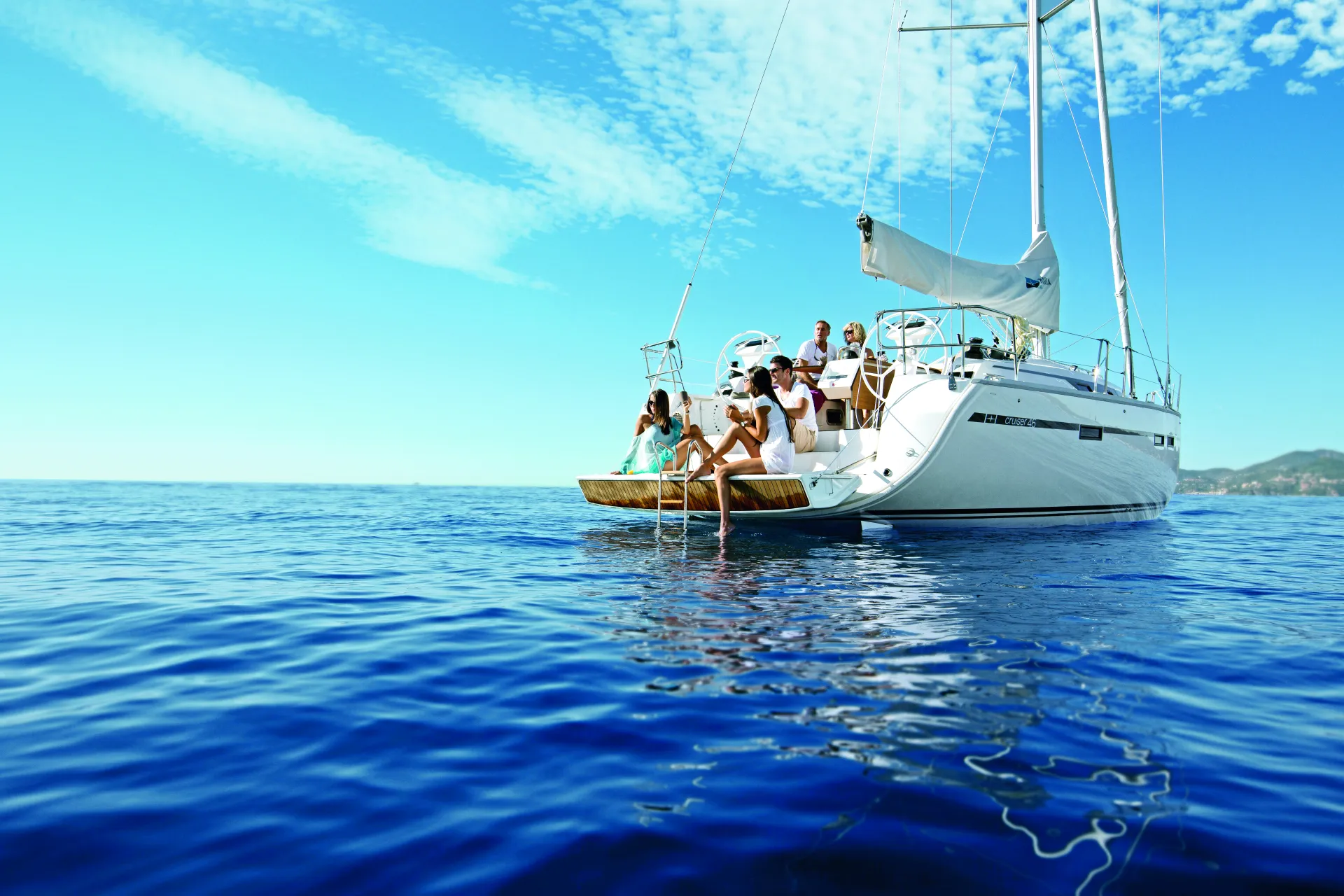 A group of people relax on a white Bavaria 46 Style sailboat in calm, clear blue water under a bright sky with scattered clouds. Some sit at the back, dipping their feet into the sea. A coastline is visible in the distance.