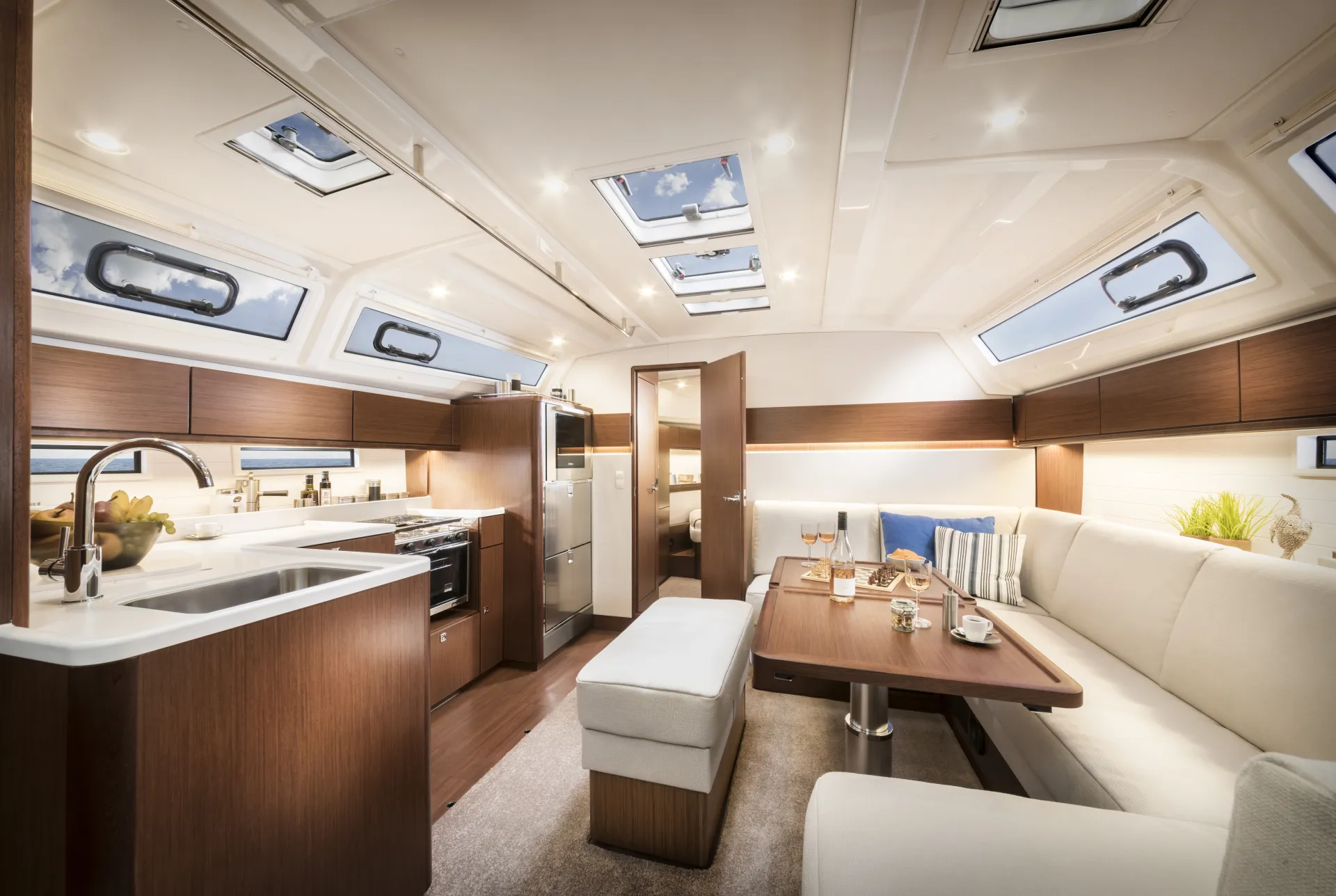 Modern Bavaria 46 Style yacht interior with wood accents, white worktops, kitchen area, lounge with beige seating, dining table set for two, and large windows letting in natural light; bright and cosy atmosphere.