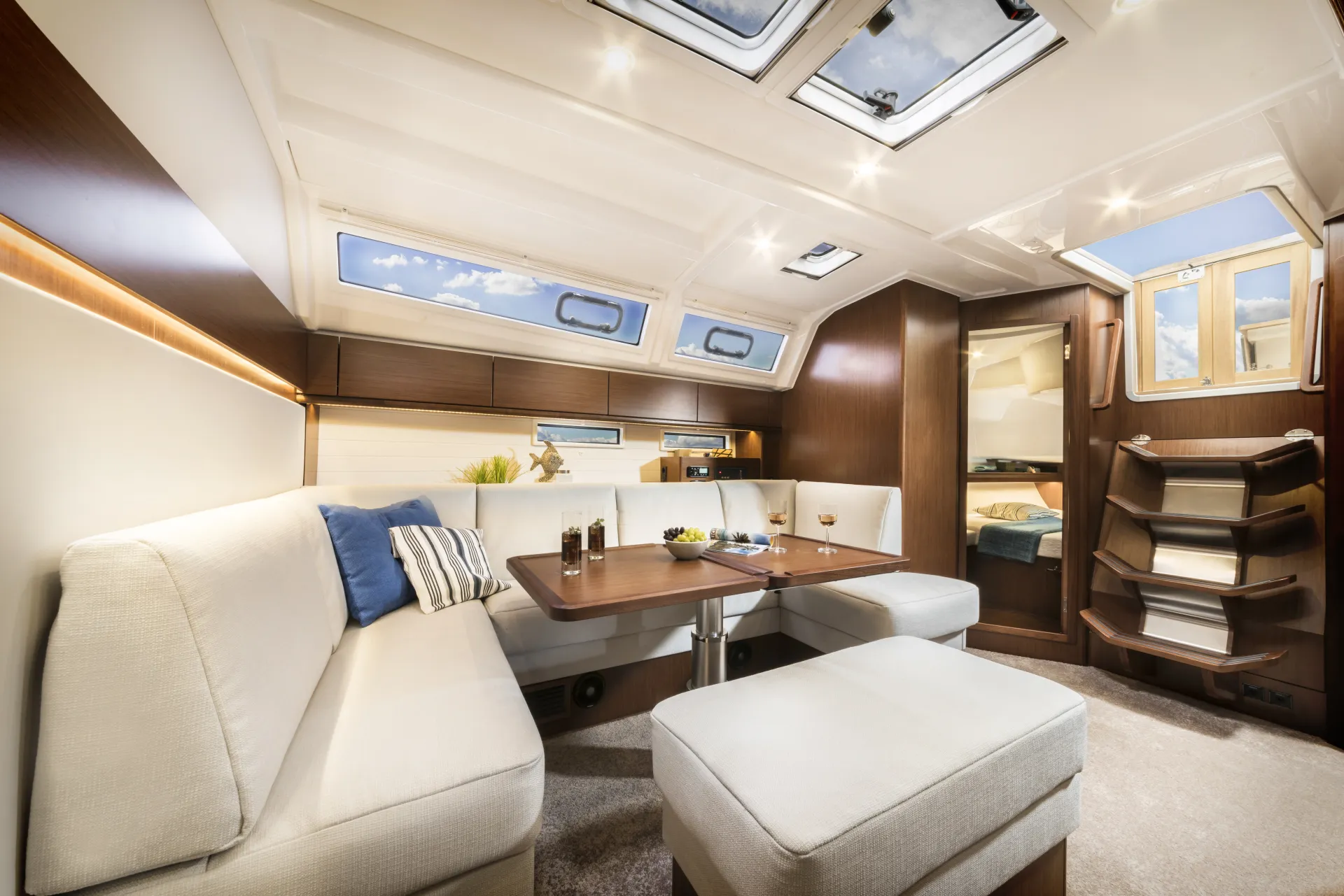Modern yacht interior aboard the Bavaria 46 Style, featuring cream sofas, wood-panelled walls, a dining table set with drinks, and skylights above. A small bedroom is visible through an open doorway and stairs lead up to a sunlit exit.