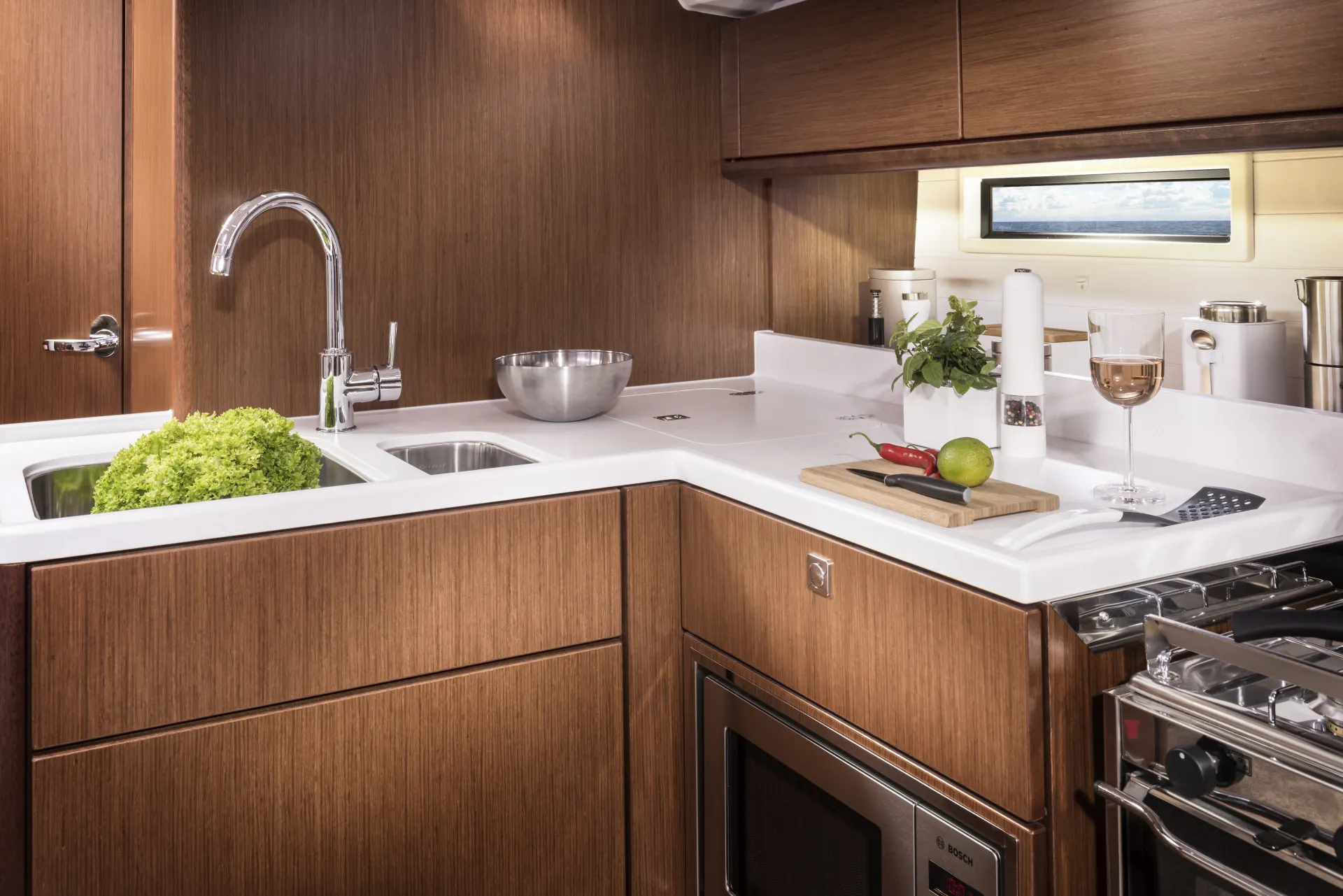 Modern kitchen with wooden cupboards, a sink, a metal bowl, lettuce, herbs, a chopping board with vegetables, a glass of wine, and a cooker with an oven—designed in the elegant style of the Bavaria 46 Style yacht. Natural light comes in through a small window.