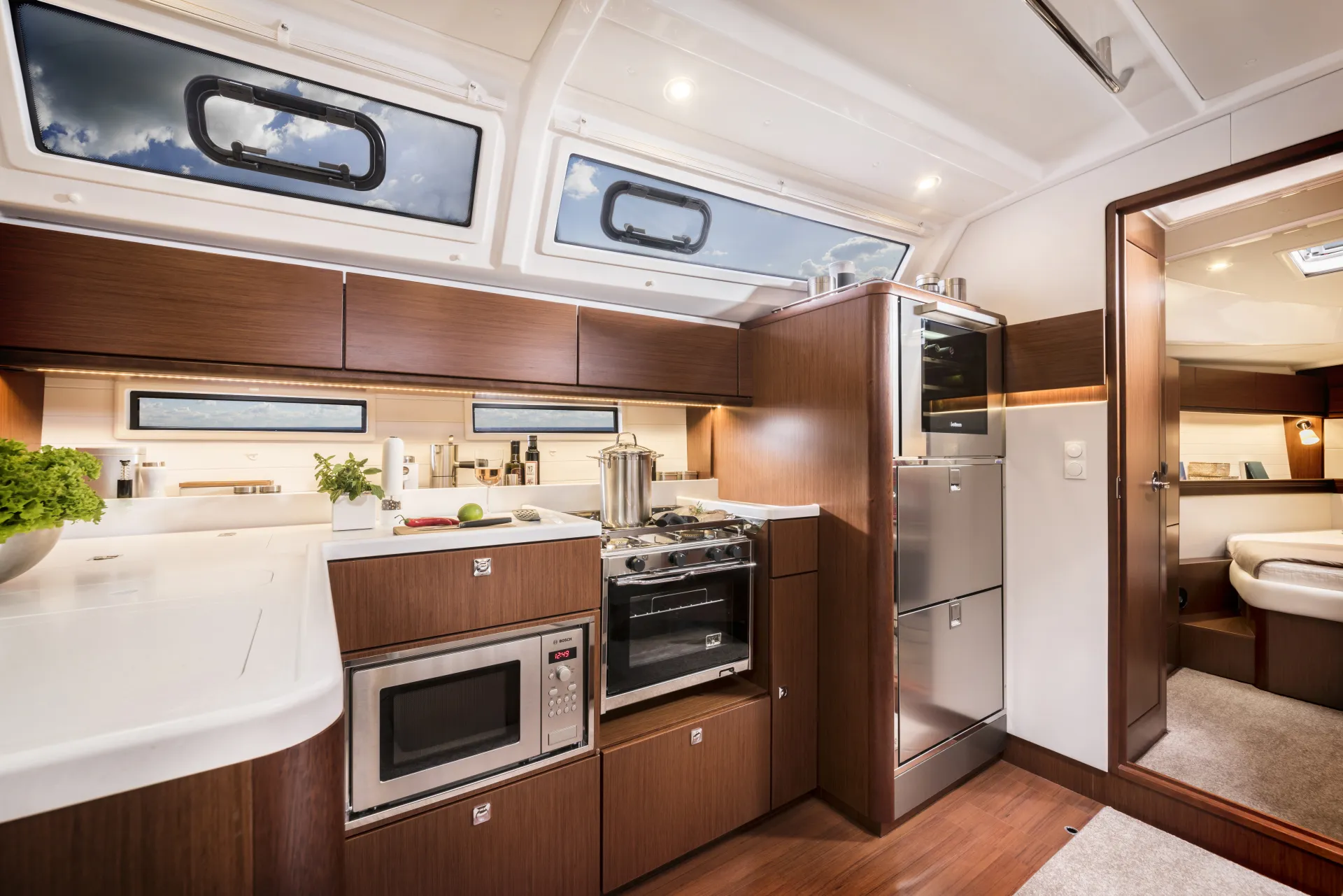 Modern yacht galley aboard the Bavaria 46 Style, with wooden cupboards, stainless steel appliances including a microwave and oven, and large windows above. Fresh herbs and vegetables are on the white worktop. A cosy bedroom is visible through an open doorway.