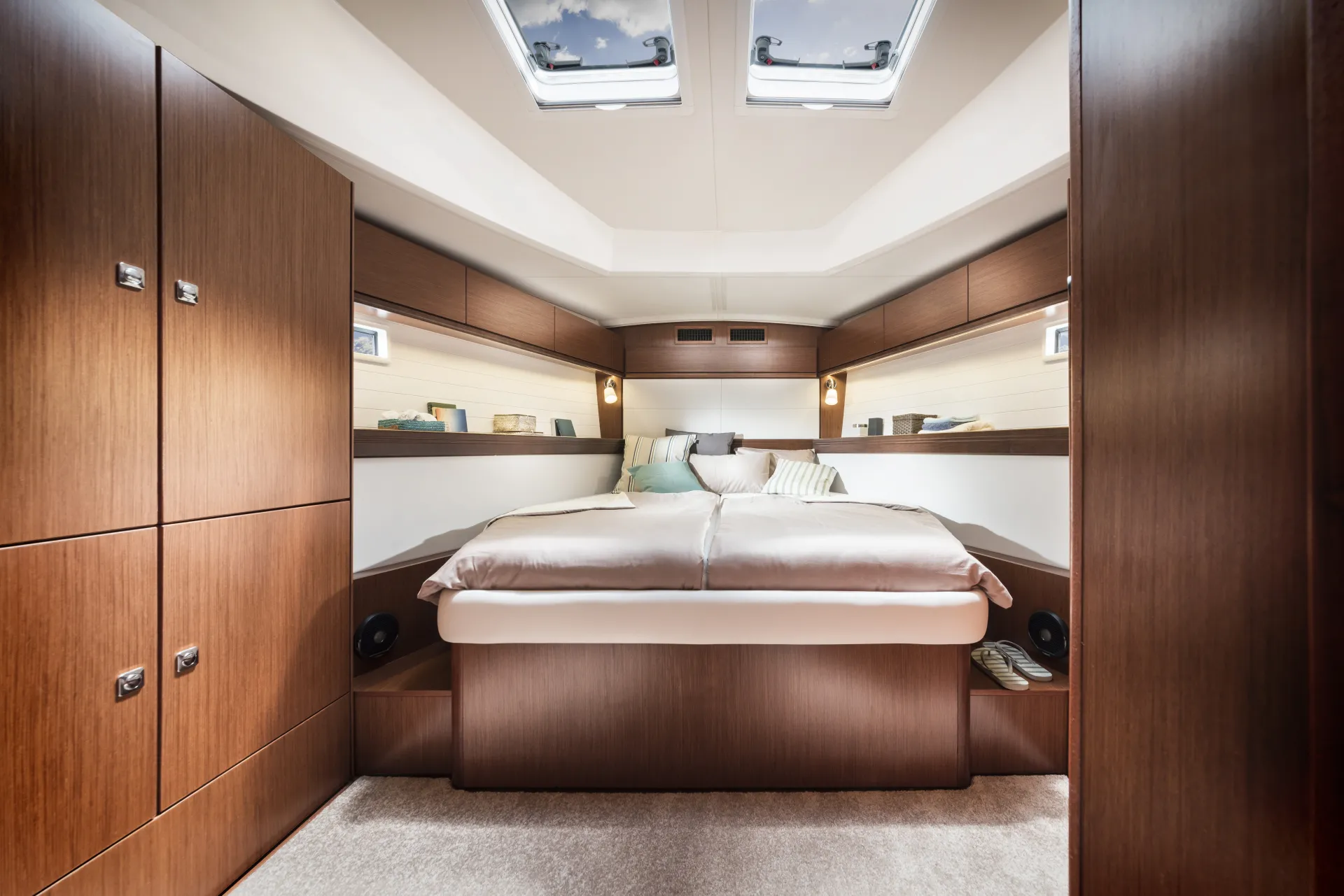 A modern yacht cabin aboard the Bavaria 46 Style features a large double bed, wood-panelled walls, built-in cupboards, reading lights, pillows, and skylights above, creating a cosy and elegant atmosphere.