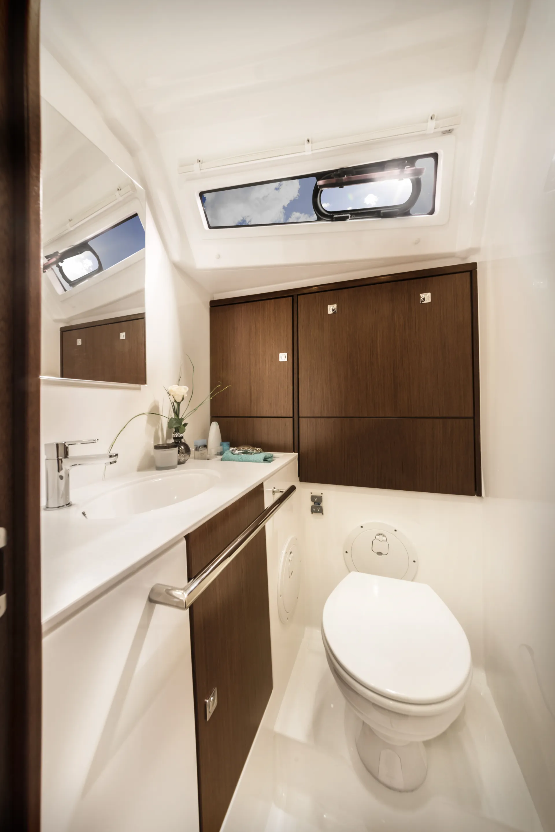 Modern yacht bathroom aboard the Bavaria 46 Style, featuring a white toilet, basin, wooden cupboards, mirror, a small plant, and a ceiling window revealing blue sky and clouds. The space is clean and compact.