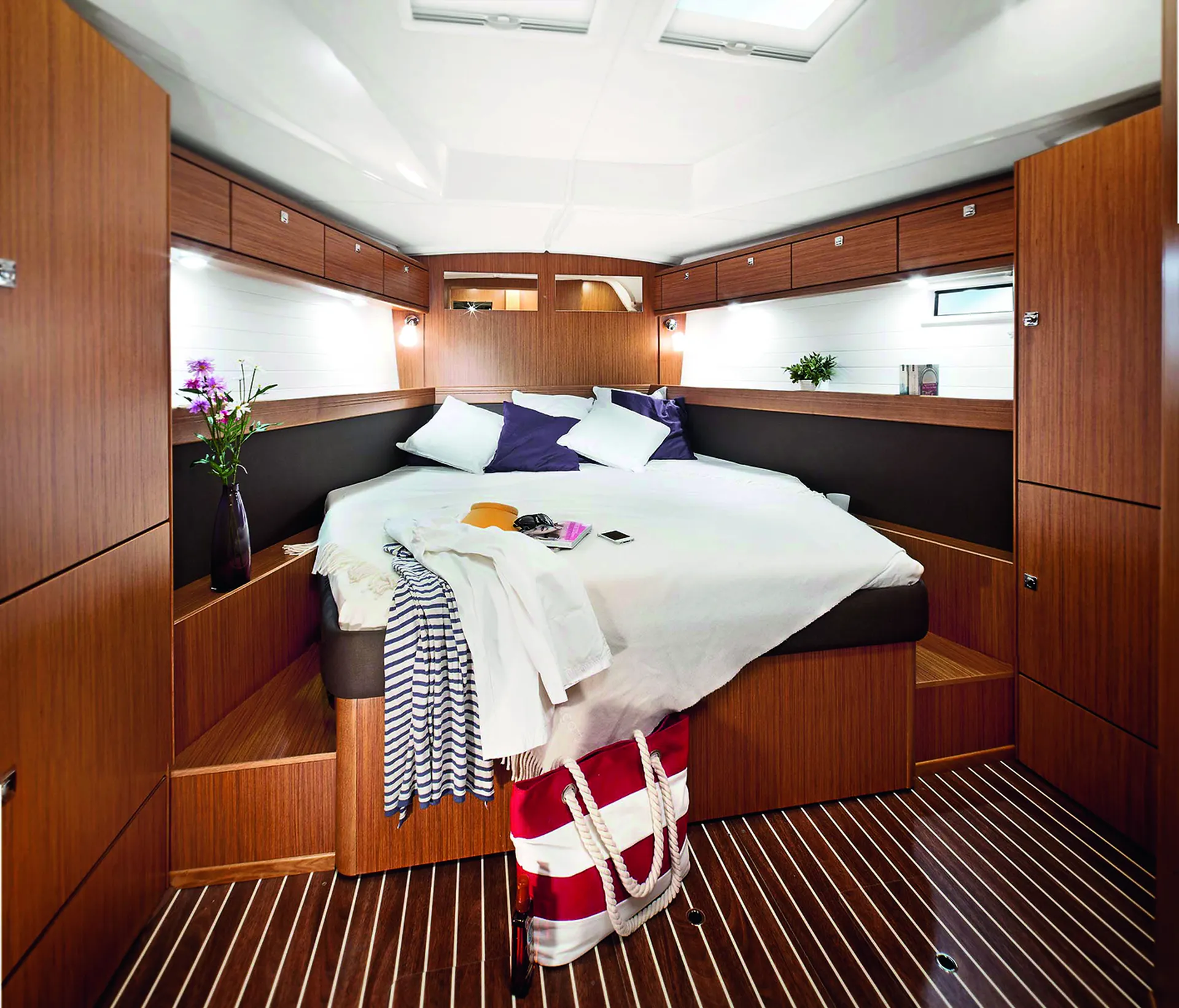 Modern boat cabin aboard a Bavaria 46 Style, with wood panelling, a large bed with white bedding and pillows, clothes and a bag on the bed, flowers in vases, built-in cupboards, and striped wood flooring.