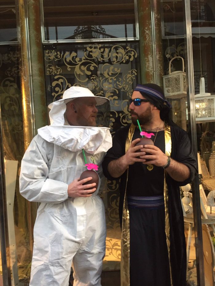 Two men in costume holding drinks and conversing. one is dressed in a white hazmat suit, and the other wears a middle eastern outfit with sunglasses. they stand in front of a decorative wall.