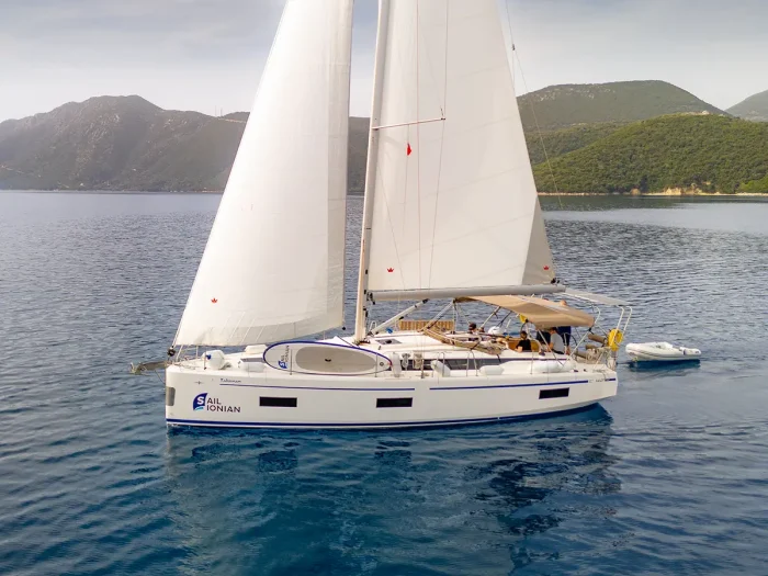 A white sailboat, the Bavaria C42, with two large sails glides on calm, clear blue water. The boat has a covered cockpit area, and the name 