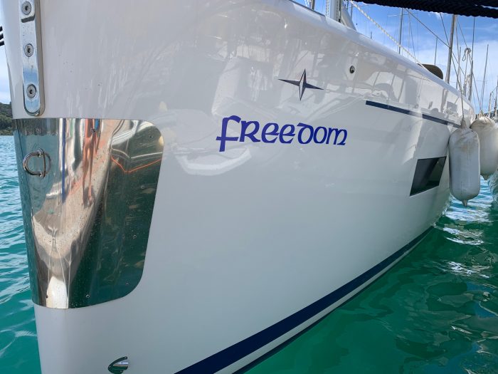 Side view of a white Bavaria C42 yacht named "Freedom" docked in a marina, showing part of its reflective hull and the blue water around it.