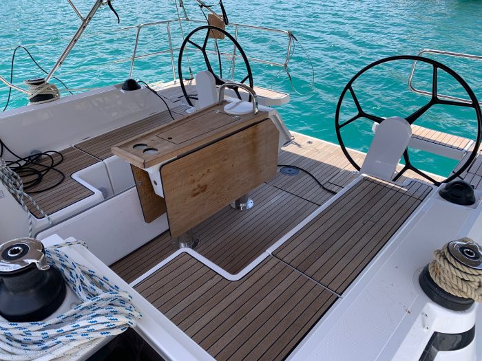 Rear deck of a Bavaria C42 sailboat showing steering wheels, a wooden table, white seats, and ocean view. The deck is equipped with various sailing gear and ropes, with clear blue