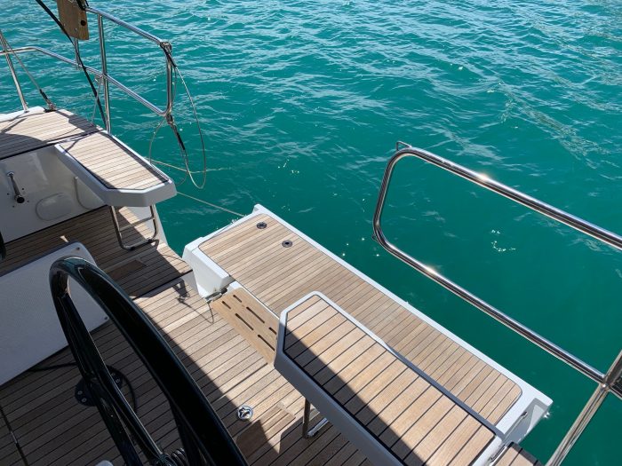 View from the aft deck of a Bavaria C42 yacht showing wooden flooring, a steering wheel, and clear turquoise water. Bright sunny day with calm sea conditions.