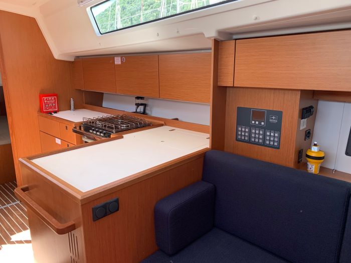 Interior of a modern Bavaria C42 yacht galley with wooden cabinets, a stove, countertop, and a blue sofa, showcasing a compact and functional design with natural light.