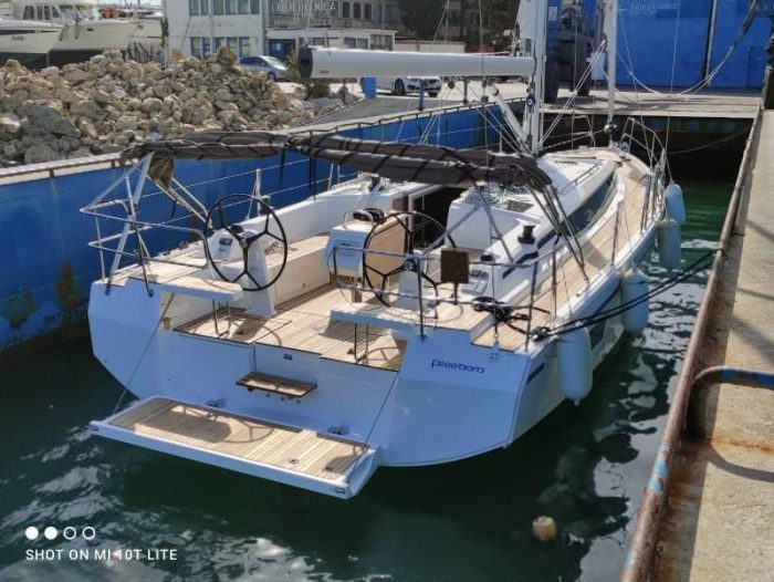 A Bavaria C42 sailboat docked at a marina with clear blue skies in the background. The boat features a dual steering wheel and a spacious deck.
