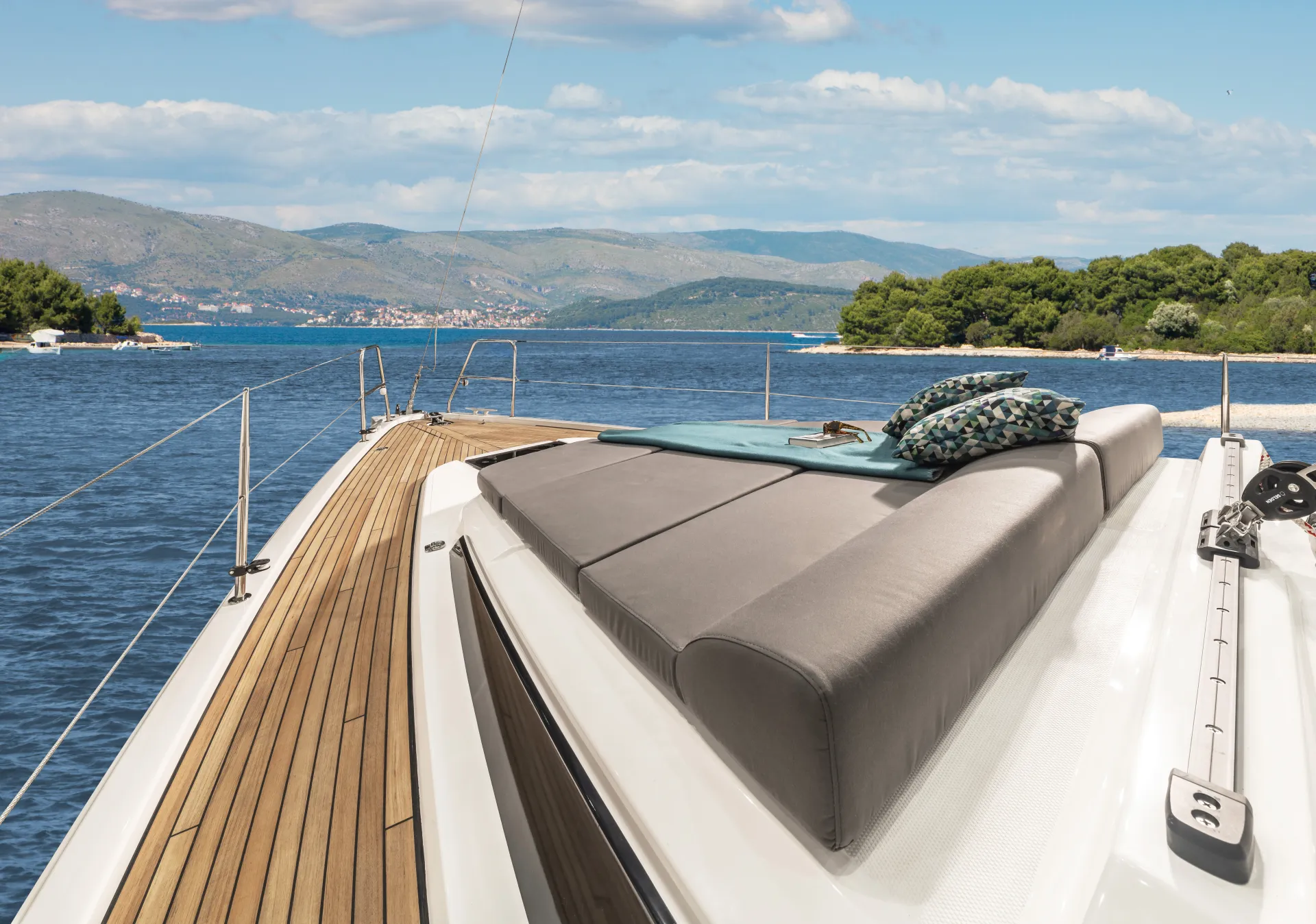A yacht deck aboard the Bavaria C42 with cushioned sunbeds and pillows, floating on calm blue water near a lush green shoreline under a partly cloudy sky.