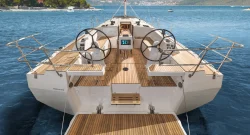 View of a modern Bavaria C42 sailboat’s teak deck from the stern, showing dual steering wheels, navigation panels, and the open sea ahead with a coastline and distant buildings visible in the background.