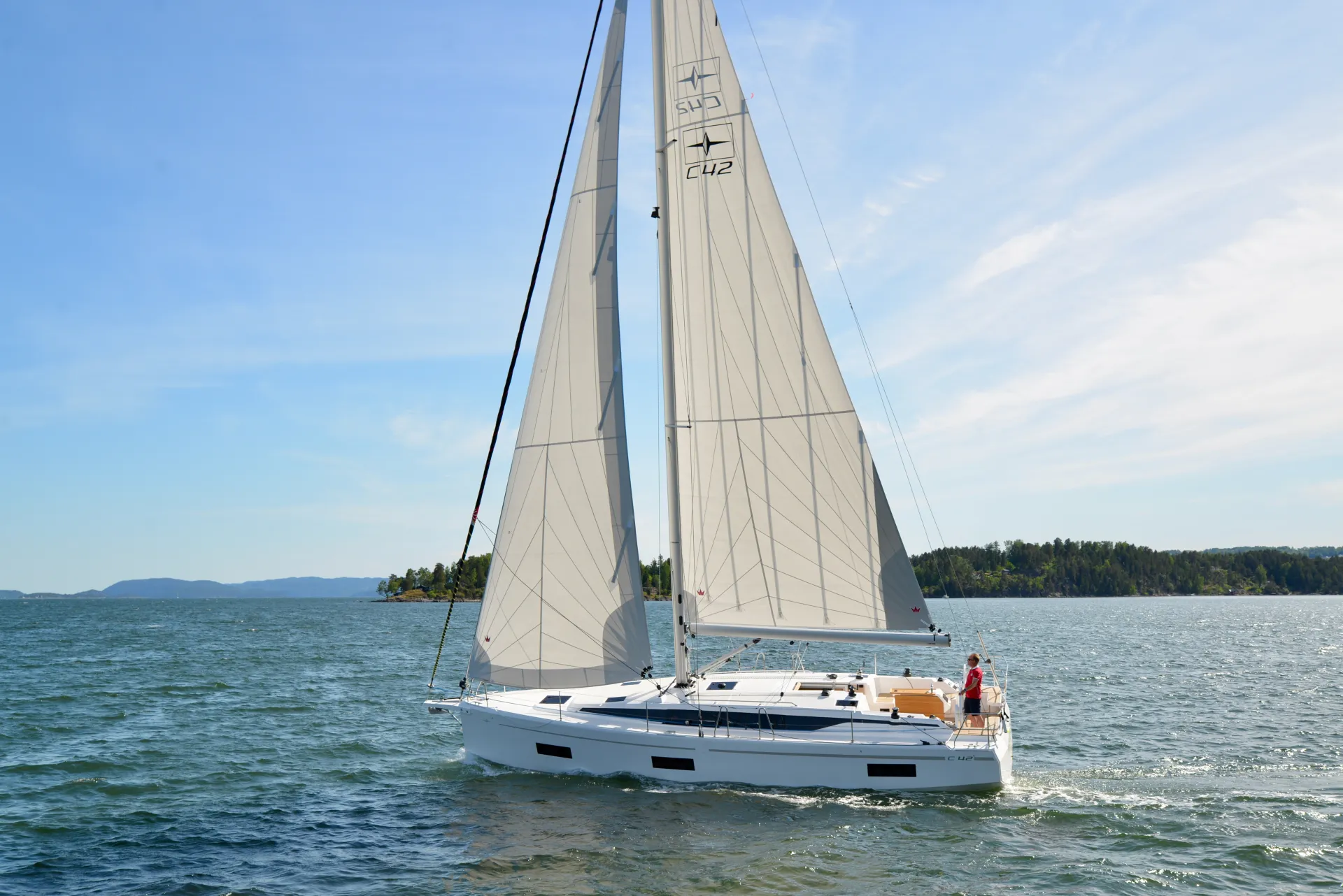 A white Bavaria C42 sailboat with raised sails glides on blue water near a forested shoreline under a clear sky. One person stands at the back of the boat, steering.