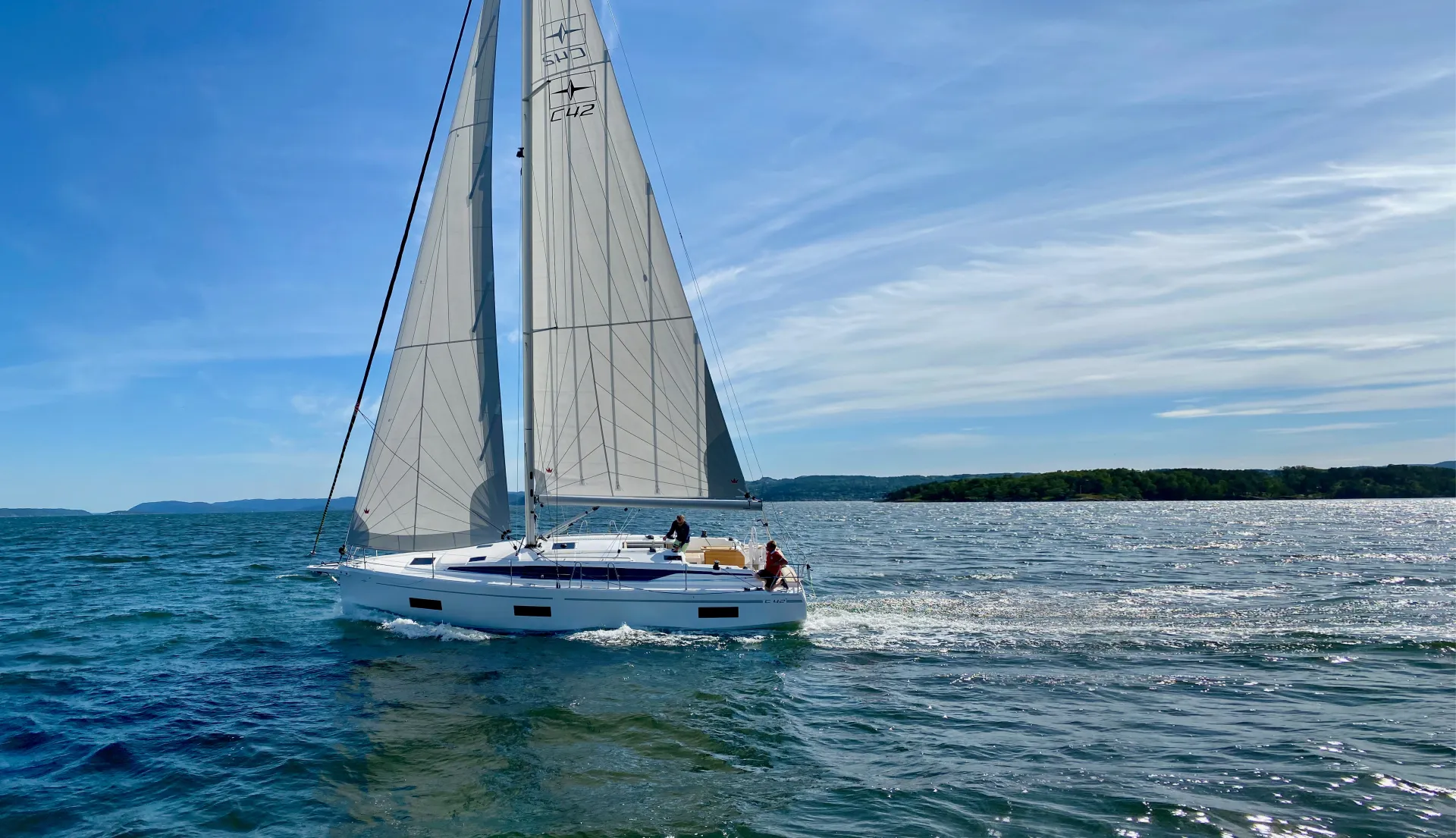 A white Bavaria C42 sailboat with raised sails glides on a blue lake under a sunny sky, with two people on deck. There are distant green trees and low hills along the shore in the background.