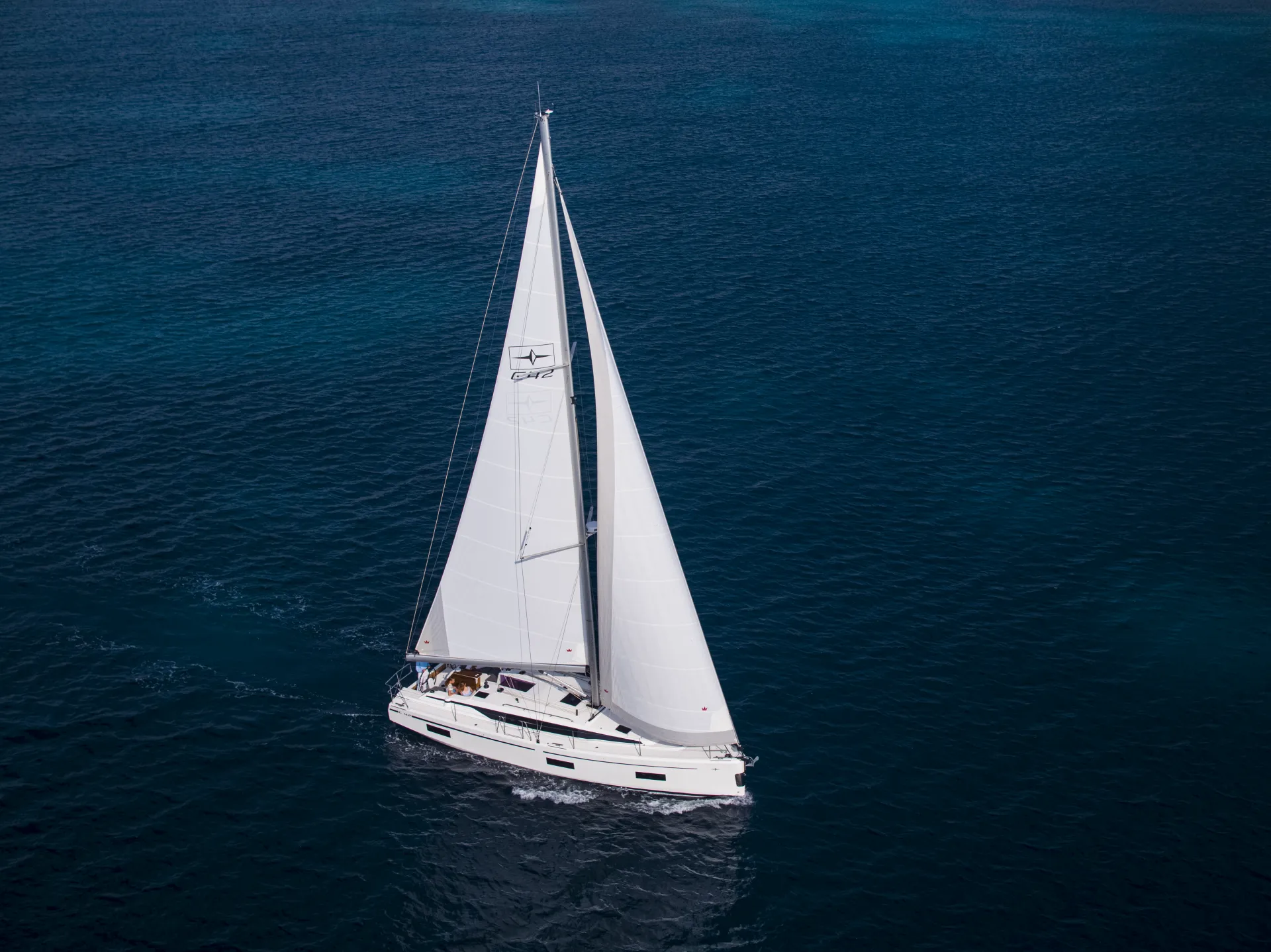 A white Bavaria C42 sailboat with large sails glides on deep blue ocean water, creating gentle ripples. The boat is seen from above under clear skies.