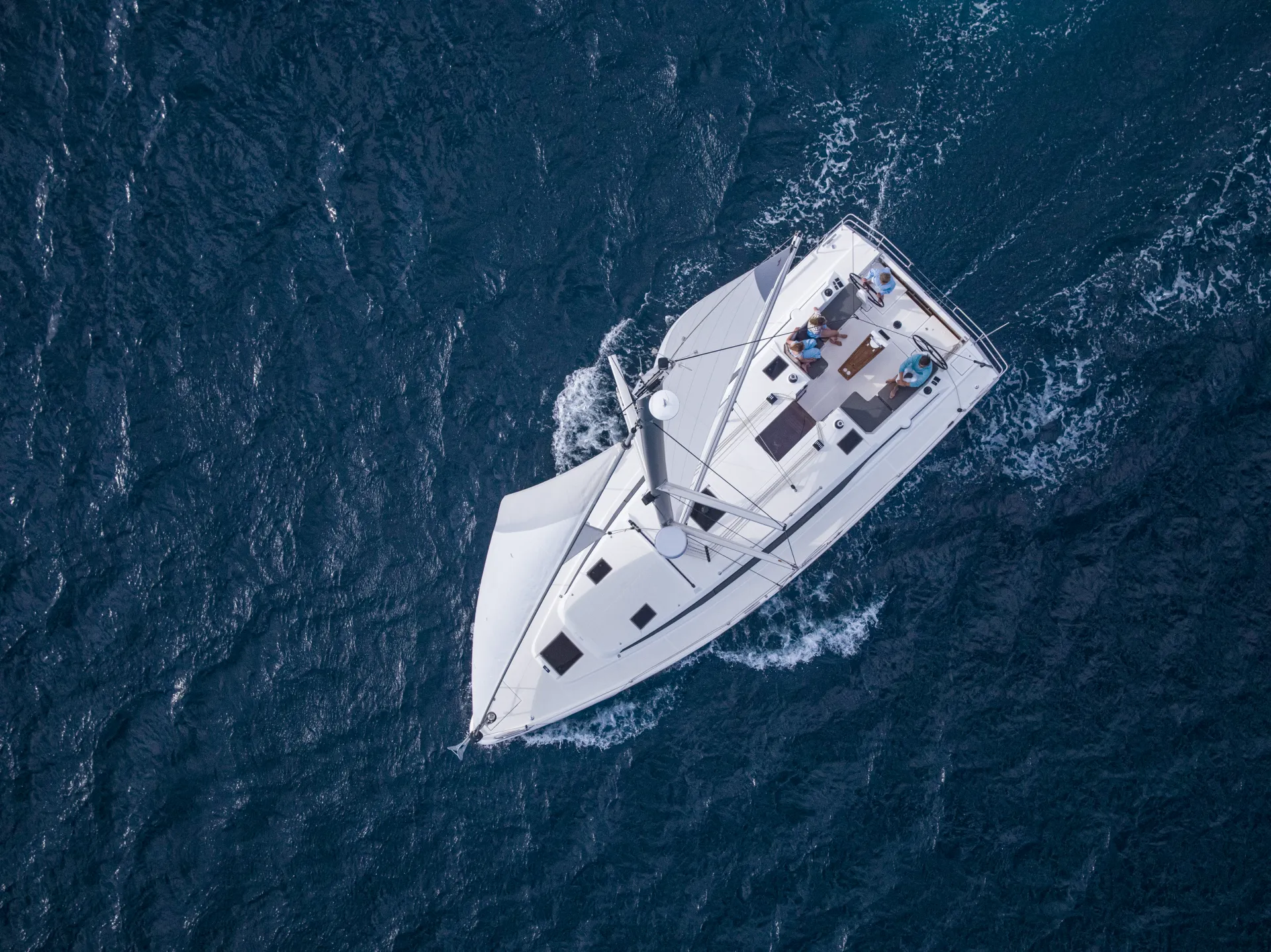 Aerial view of a white Bavaria C42 sailboat sailing on deep blue water, with visible waves and two people relaxing on the deck.