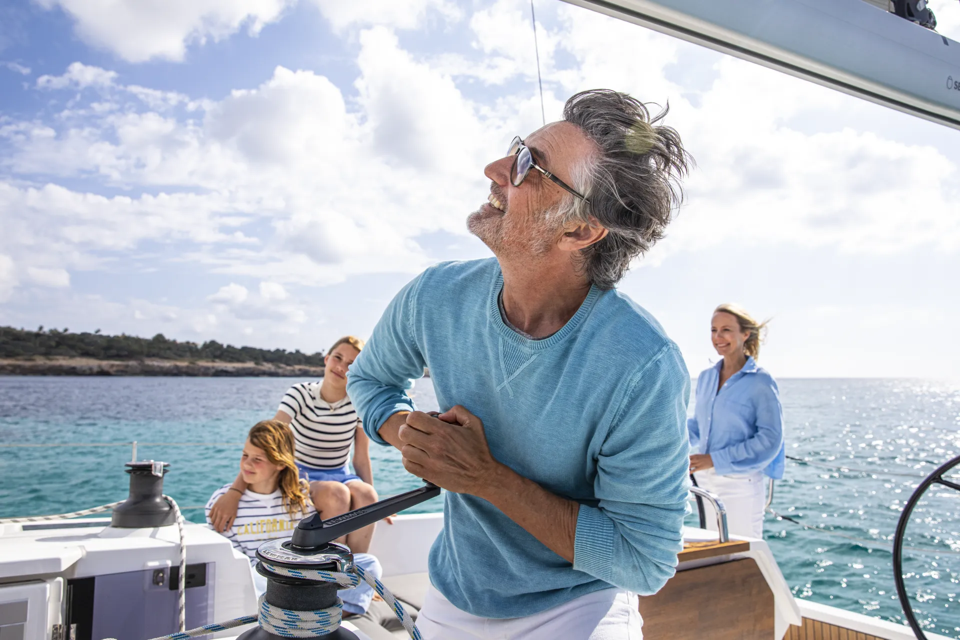 A man in sunglasses sails a Bavaria C42, turning a winch while three smiling people relax on board. The sun is shining and the sea is calm in the background.