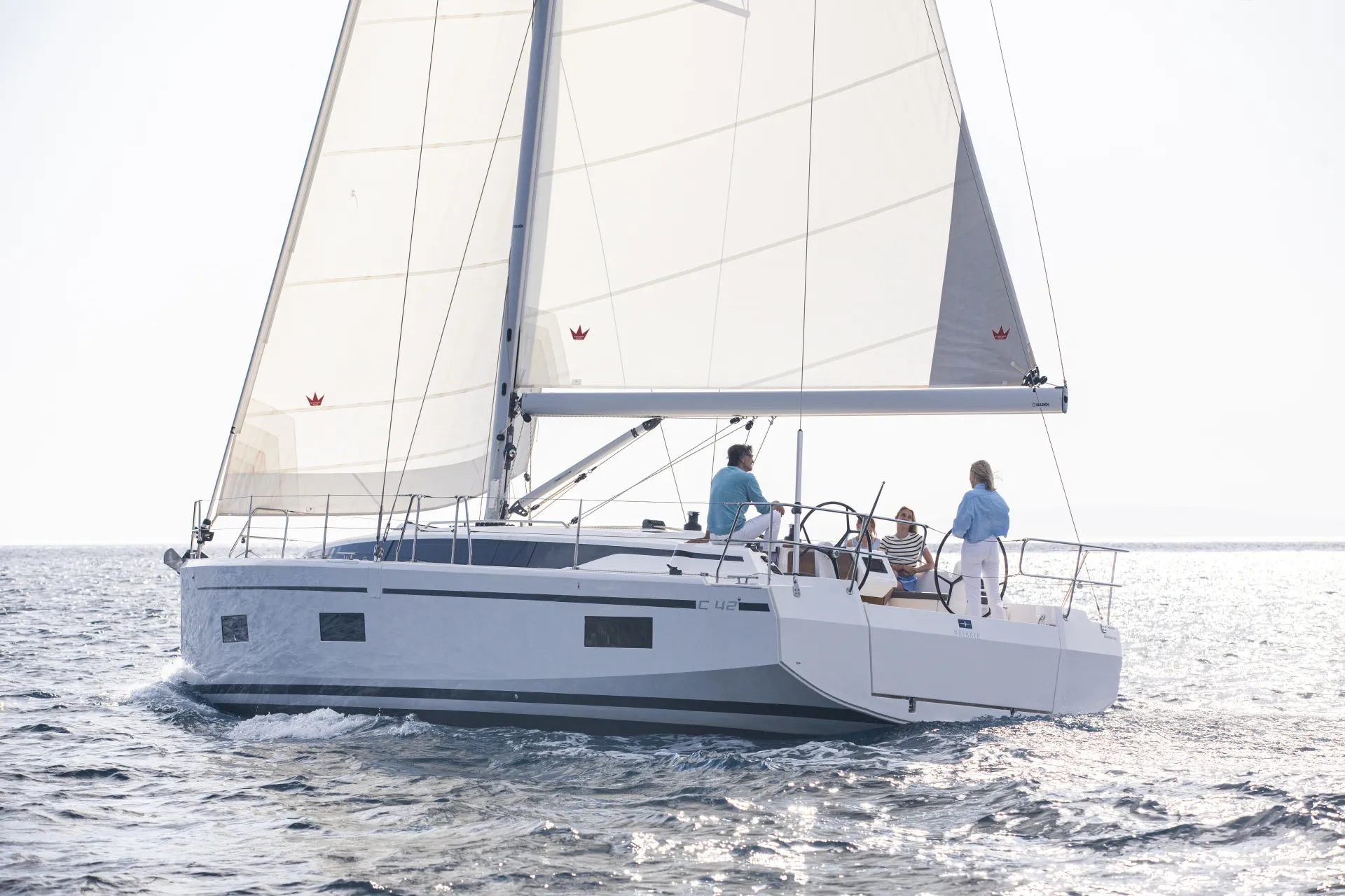 Three people sail a modern white Bavaria C42 yacht on calm, open water under clear skies. The sails are full, and the boat moves smoothly, creating small waves. The group appears relaxed, enjoying the sunny day.