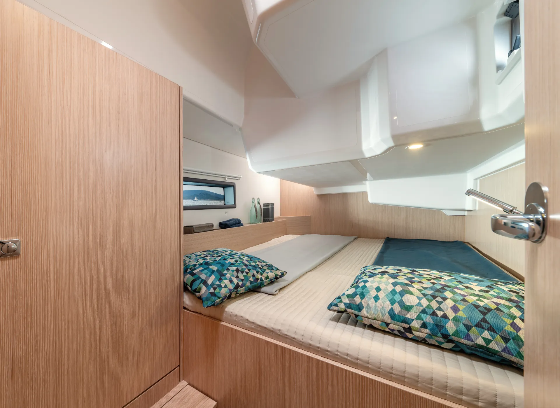 A compact, modern boat cabin aboard the Bavaria C42 features wooden walls, a large bed, two geometric-patterned cushions, and a window letting in natural light.
