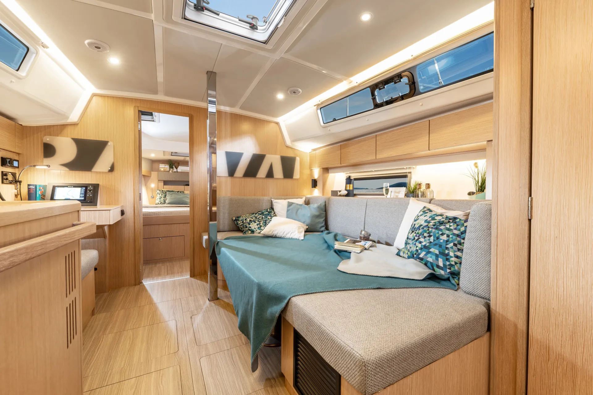 Modern yacht interior with light wood finishes, a grey sofa with blue and patterned cushions, a small table with books, and large windows providing natural light. The space looks bright, cosy, and organised—an inviting retreat that perfectly reflects the elegant design of the Bavaria C42.
