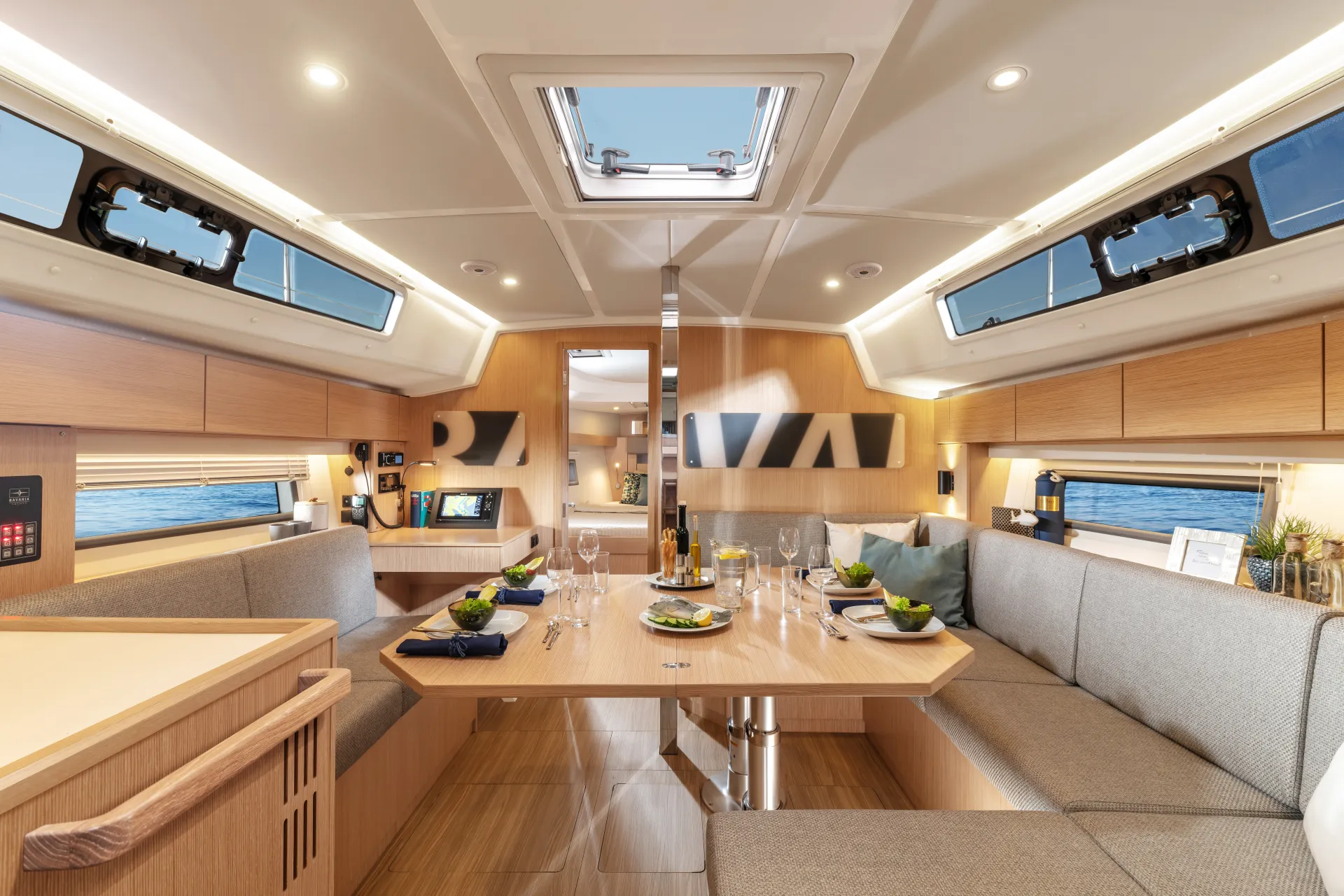 Modern yacht interior with light wood, large windows, grey cushioned seating, a dining table set with plates and glasses, and ocean visible outside. The inviting atmosphere of the Bavaria C42 is bright and clean, perfect for relaxing or entertaining at sea.
