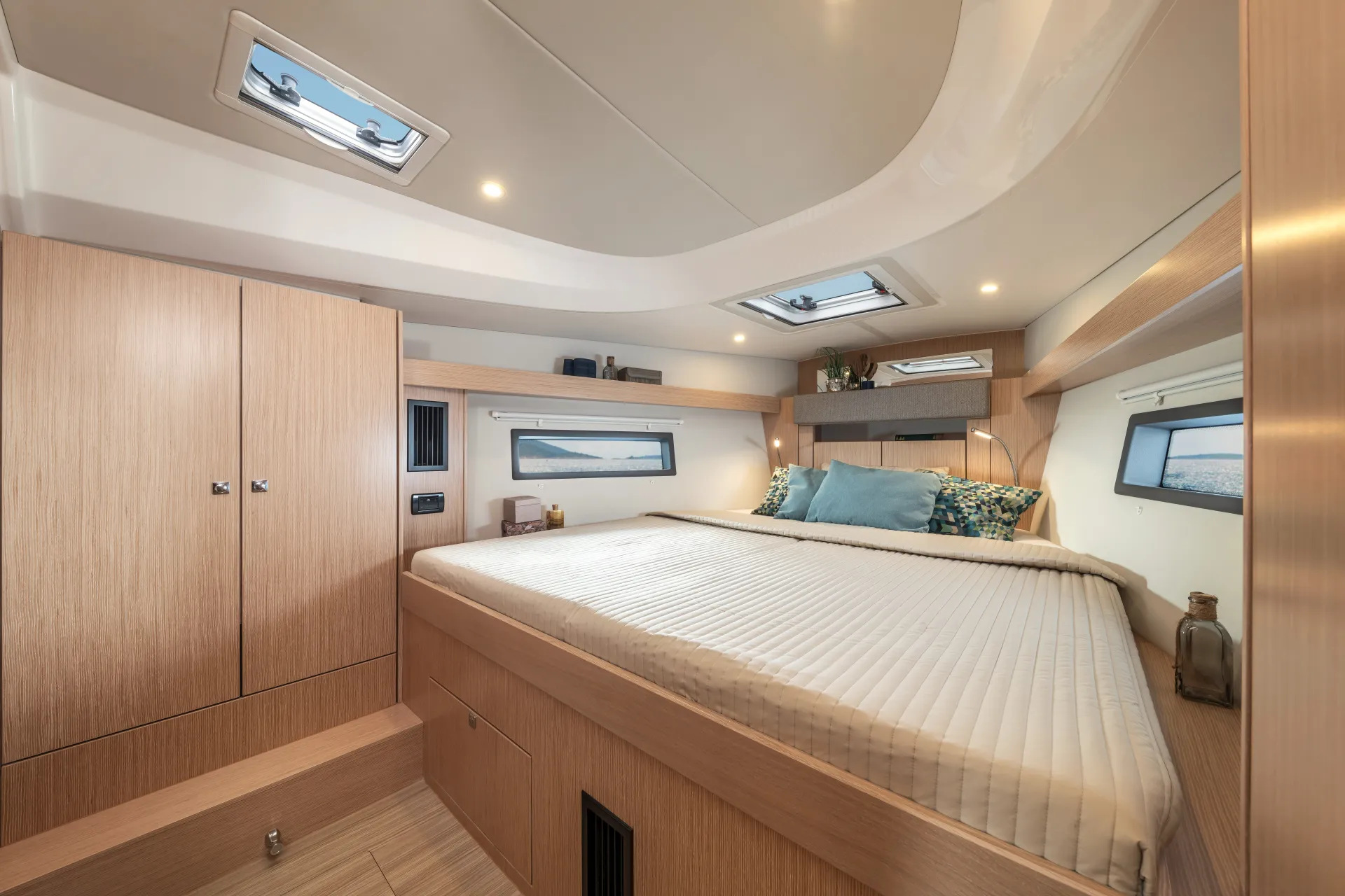 Modern boat cabin bedroom aboard the Bavaria C42, featuring a large bed, wooden cupboards, bedding with pillows, windows on the ceiling and sides, and soft lighting. A bottle and small décor items are arranged on shelves beside the bed.
