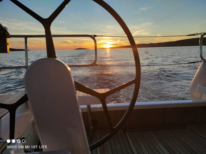 A sunset view from the deck of a boat in Vliho, showing a steering wheel in the foreground, with the sun setting over the horizon and the sea in the background.
