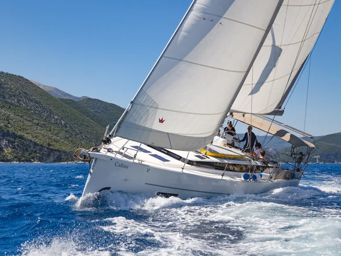 A Bavaria C45 glides through bright blue water, sails fully extended, with several people on board, against green hills and a clear sky—a perfect scene for a luxury yacht charter.