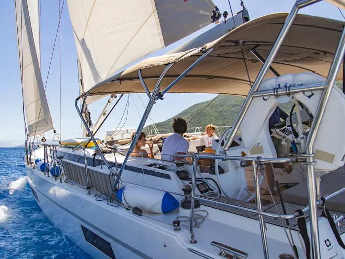 A group of people relax on the deck of a Bavaria C45, enjoying a luxury yacht charter as they cruise over blue water with mountains in the background beneath a clear sky.