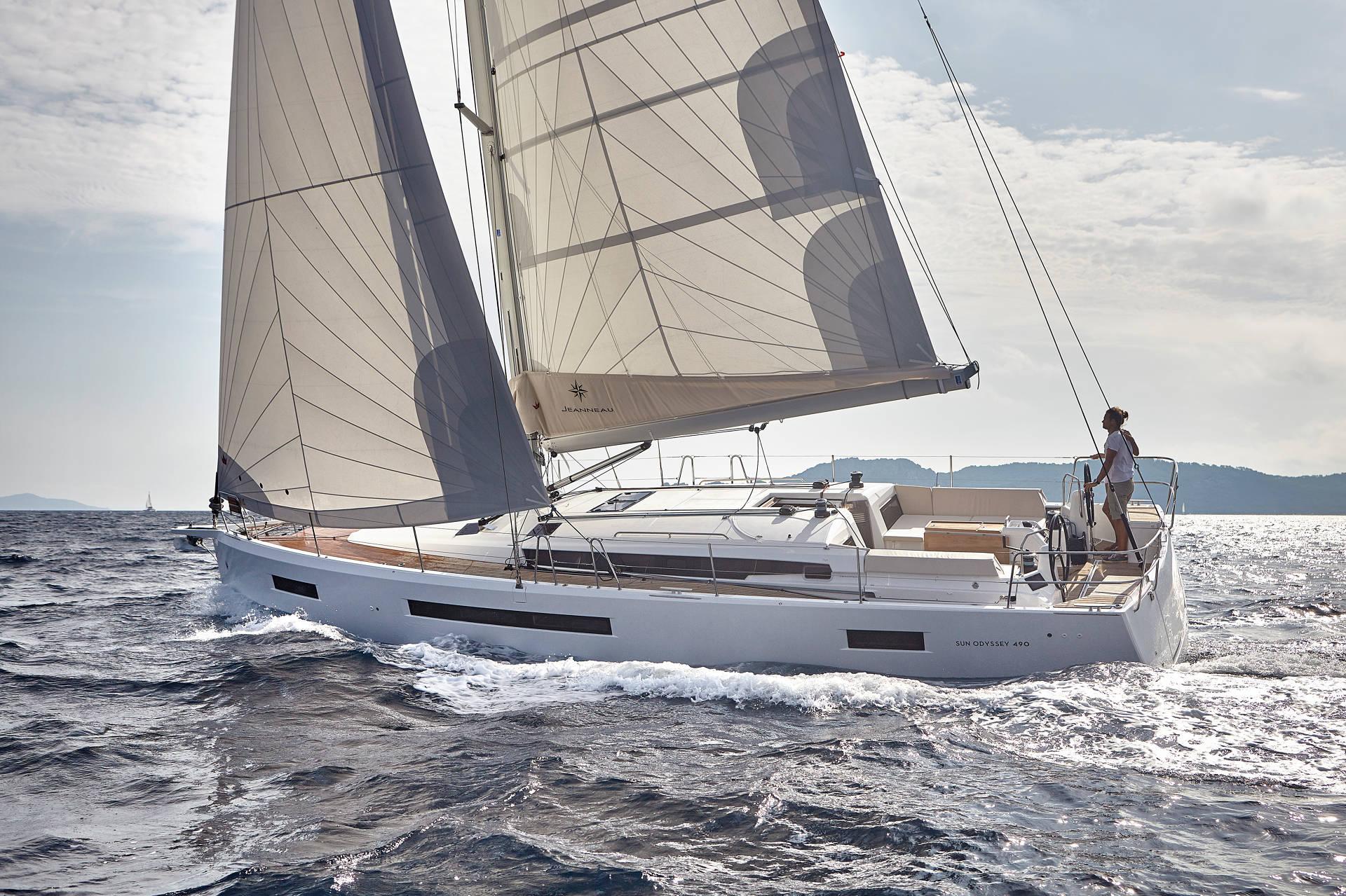 A sailboat with large white sails glides through the ocean during the 2022 sailing season, with a person standing at the helm, steering. The horizon is lined with distant mountains under a mostly