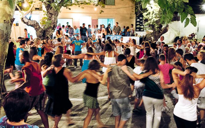 People dancing hand in hand in a circle at a lively outdoor evening event in a village square during Kalo Dekapentavgoustos, surrounded by onlookers.