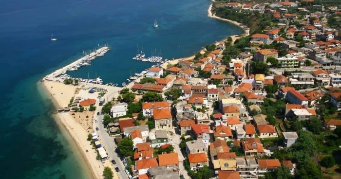 Aerial view of Palairos with tightly packed buildings, a sandy beach, and a marina with several boats. The landscape includes greenery and the sea extends into the horizon.