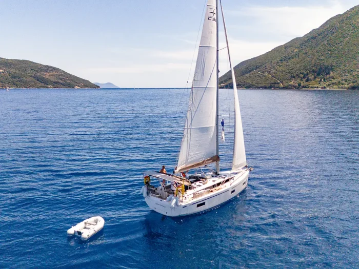 A white Bavaria C38 sailboat with its sails up glides through calm, clear blue water, followed closely by a small inflatable dinghy. The boat is surrounded by green, hilly landscapes under a clear, sunny sky. The scene is tranquil and picturesque.