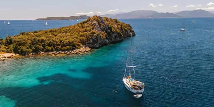 A Bavaria C38 sailboat floats on clear blue water near a small, rocky, tree-covered island, with other sailboats and distant land visible under a clear sky.
