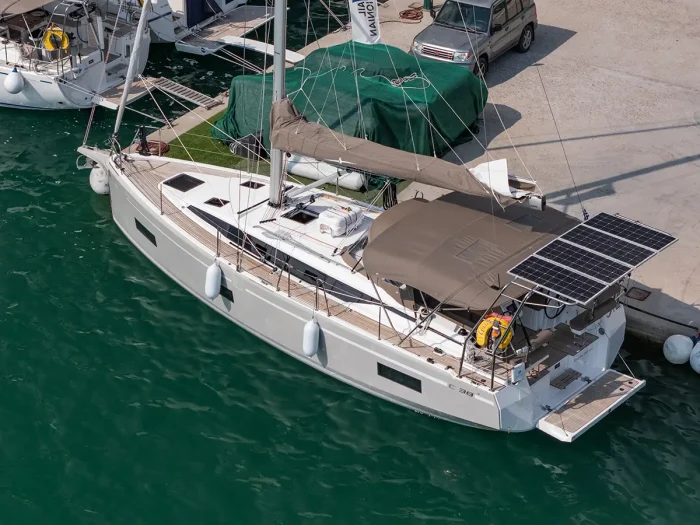 A Sail Ionian Bavaria C38 sailboat with tan coverings docked at a marina. It has solar panels mounted on the rear, fenders along the sides, and a lifebuoy attached to the back. The boat is adjacent to a concrete pier, with a car and other boats visible in the background.