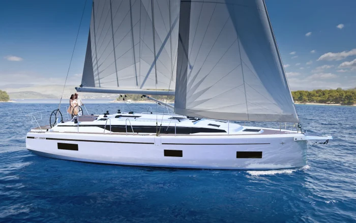 A modern white Bavaria C38 sailboat with grey sails glides over clear blue water, with two people standing on the deck. The coastline with green trees is visible in the background under a bright, cloud-dotted sky.